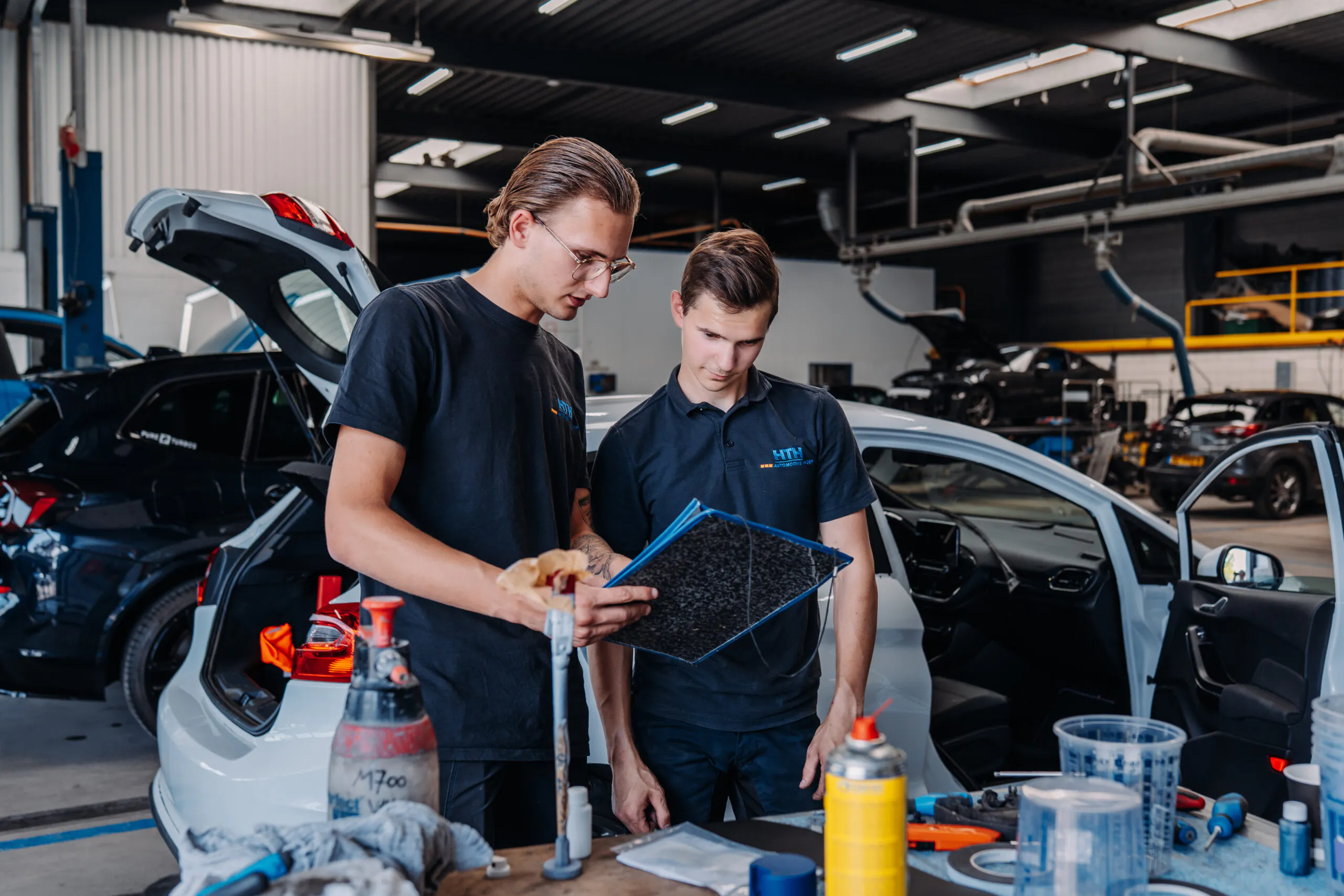 Jonge praktijkbegeleider legt een leerling een reparatietechniek uit binnen het autoschadeherstel